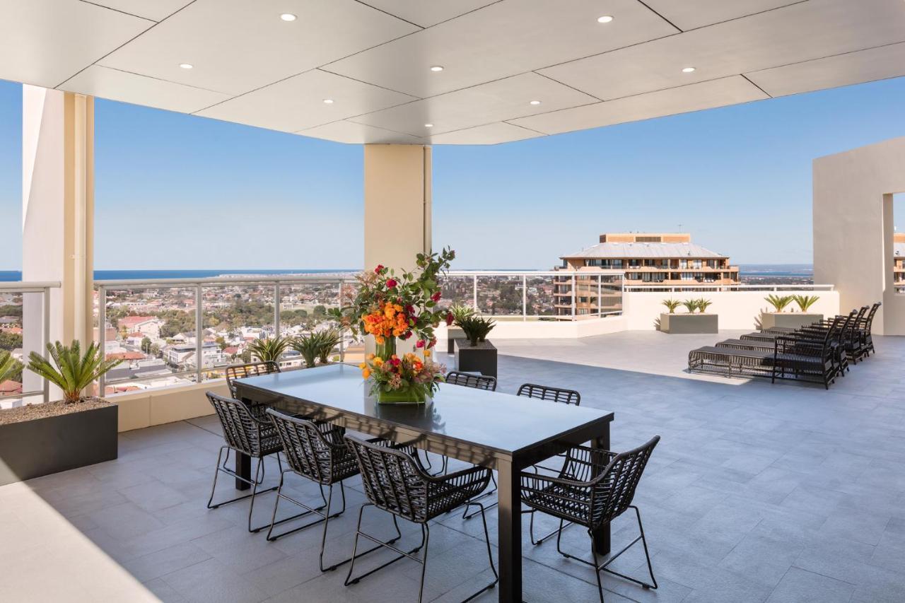 Meriton Suites Bondi Junction Sydney Exterior foto The photo shows a stylish outdoor terrace or balcony area, featuring a long dining table surrounded by several black wire chairs. The table has a centerpiece of vibrant flowers. The space is well-lit, with a clear view of the horizon in the backgroun