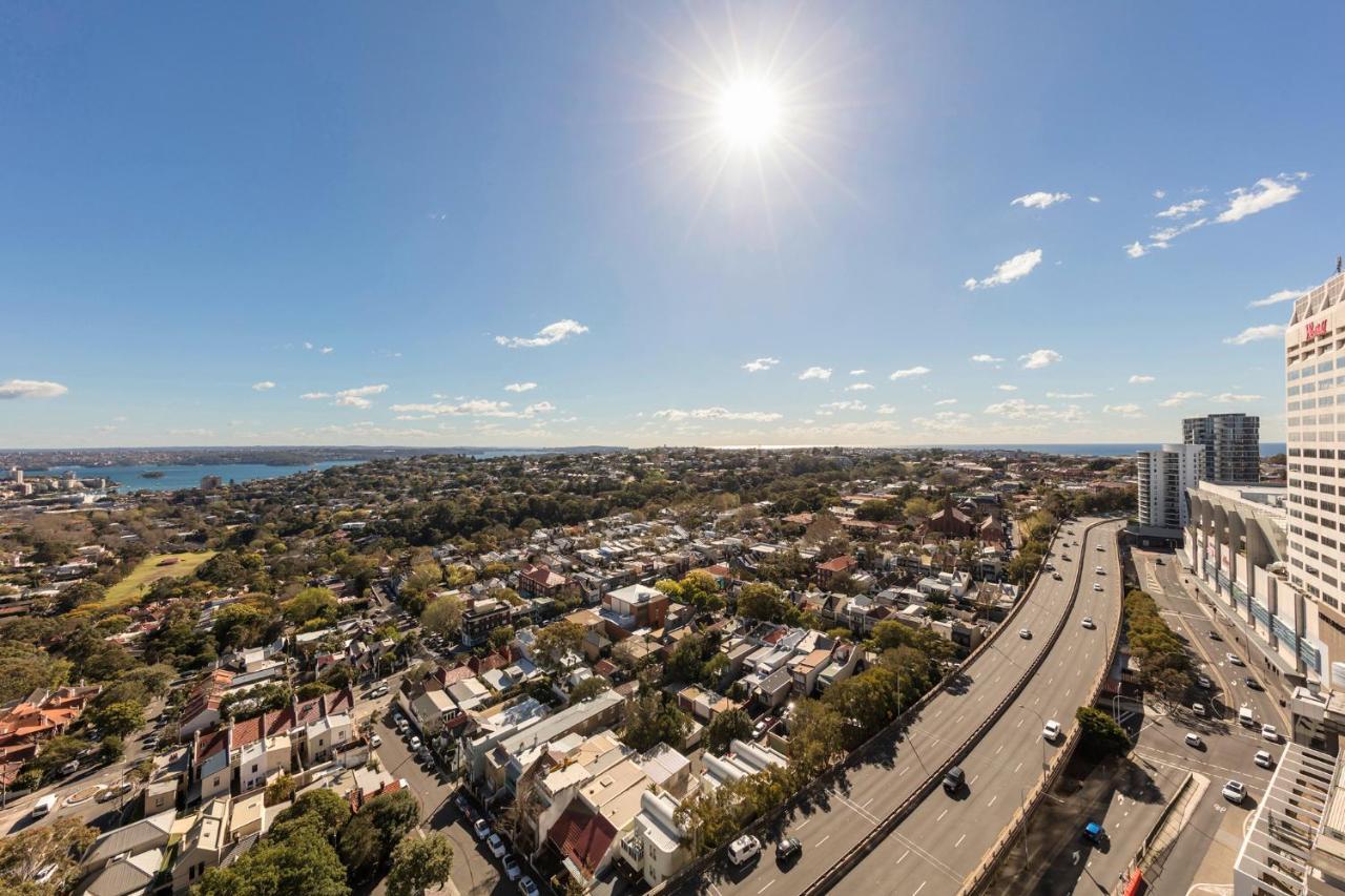 Meriton Suites Bondi Junction Sydney Exterior foto The photo shows a panoramic view of a city landscape under a clear blue sky. In the foreground, there is a road with vehicles traveling along it, bordered by trees and buildings. In the background, you can see a residential area with various houses, 