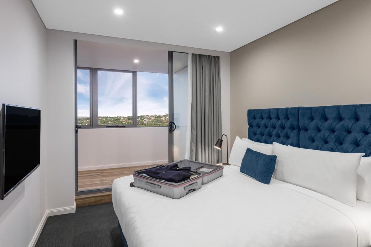 Meriton Suites Bondi Junction Sydney Exterior foto The photo shows a modern hotel room featuring a double bed with a tufted blue headboard. There is a neatly arranged suitcase placed open on the bed, indicating that someone may be unpacking. The room has light-colored walls and a window allowing natu