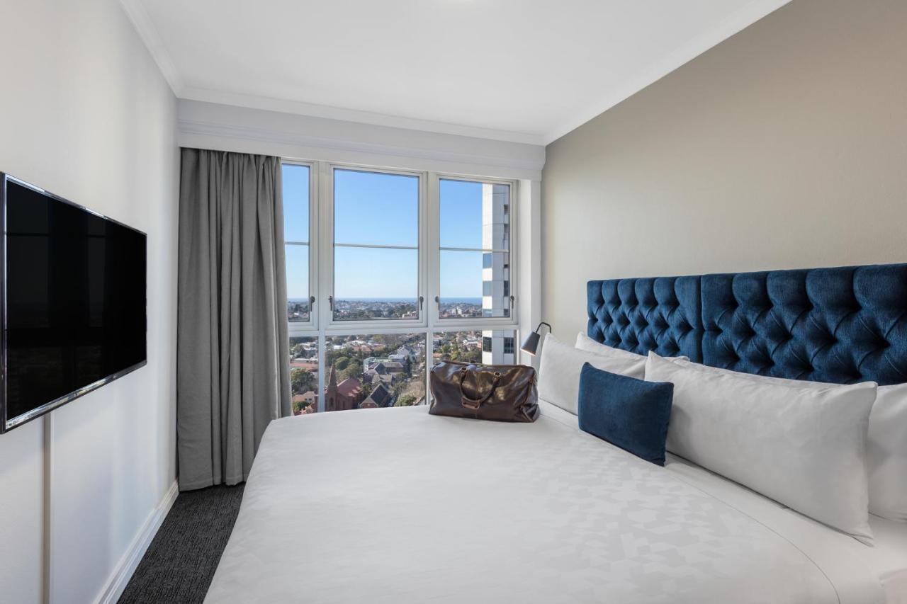 Meriton Suites Bondi Junction Sydney Exterior foto The photo shows a modern hotel room featuring a large bed with a tufted blue headboard. Beside the bed, there is a stylish bag placed on the bed. The room has large windows that provide a view of the outside, allowing natural light to fill the space.