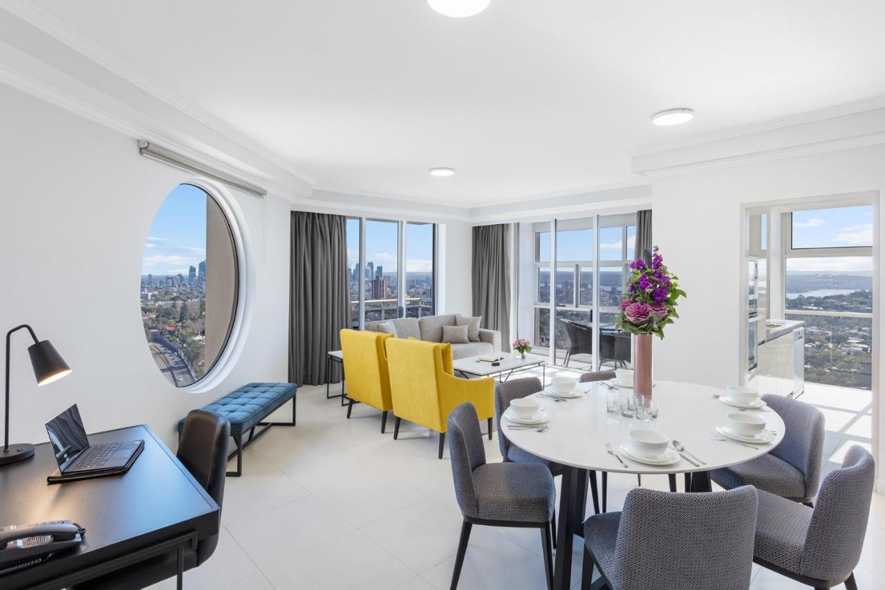 Meriton Suites Bondi Junction Sydney Exterior foto The photo shows a modern and bright interior space, likely a living room or dining area. It features large windows that offer a panoramic view of the city skyline. The furnishings include a round dining table set for several people, with plates and g