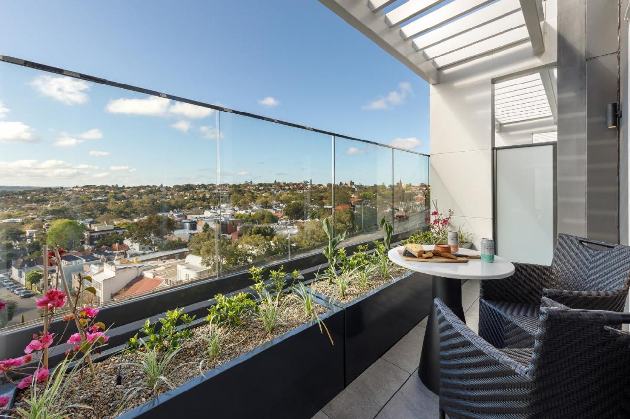 Meriton Suites Bondi Junction Sydney Exterior foto The photo shows a modern balcony with a glass railing, enhancing the view of the surrounding area, which includes various rooftops and greenery in the distance. There is a small round table set with a decorative floral arrangement and some items, lik