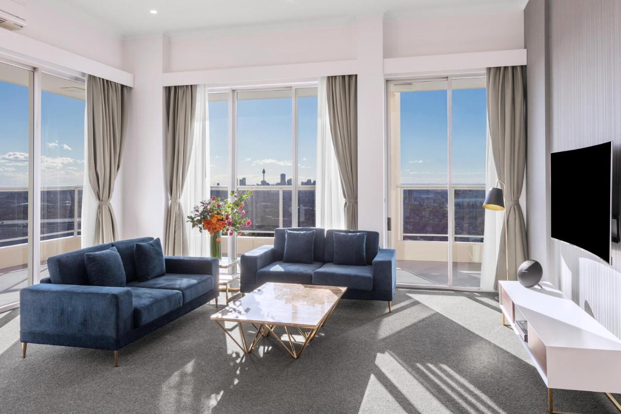 Meriton Suites Bondi Junction Sydney Exterior foto The photo shows a modern living room with large windows that allow natural light to fill the space. There are two blue sofas arranged around a coffee table with a glass top and gold legs. A floral arrangement adds a touch of color, placed on a nearby