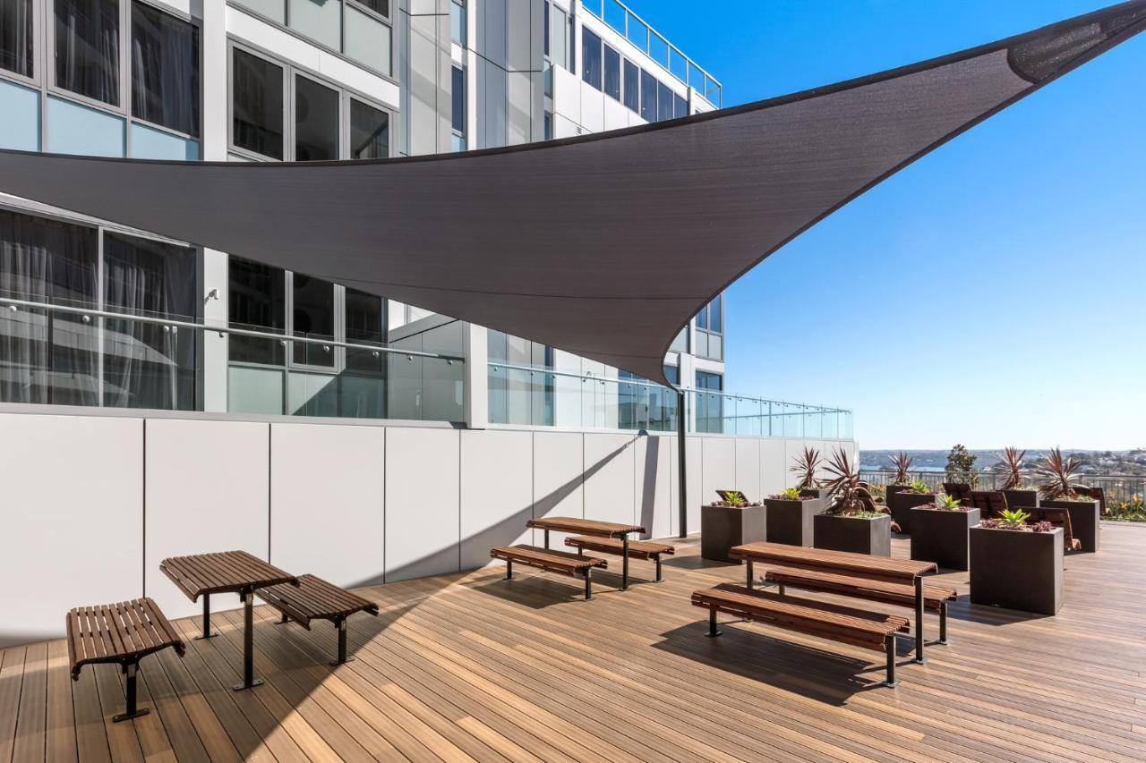 Meriton Suites Bondi Junction Sydney Exterior foto The photo shows a modern outdoor terrace area on a building. It features several wooden benches arranged on a wooden deck. There are large planters with greenery, adding some decorative elements. A large triangular gray shade sail is positioned above