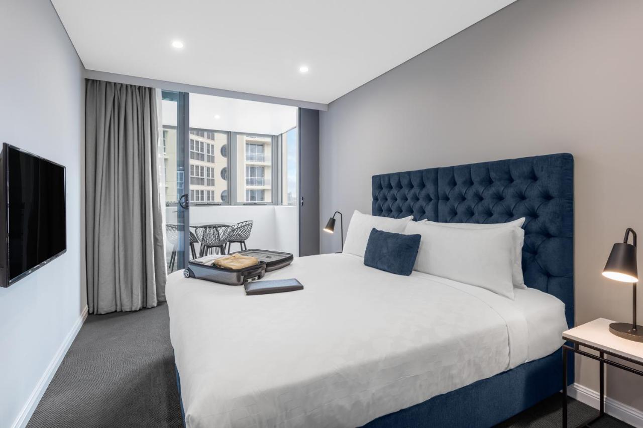 Meriton Suites Bondi Junction Sydney Exterior foto The photo shows a modern hotel room featuring a large bed with a tufted blue headboard. There are minimalistic bedside tables with lamps on either side of the bed. A flat-screen TV is mounted on the wall across from the bed. To one side, there are fl