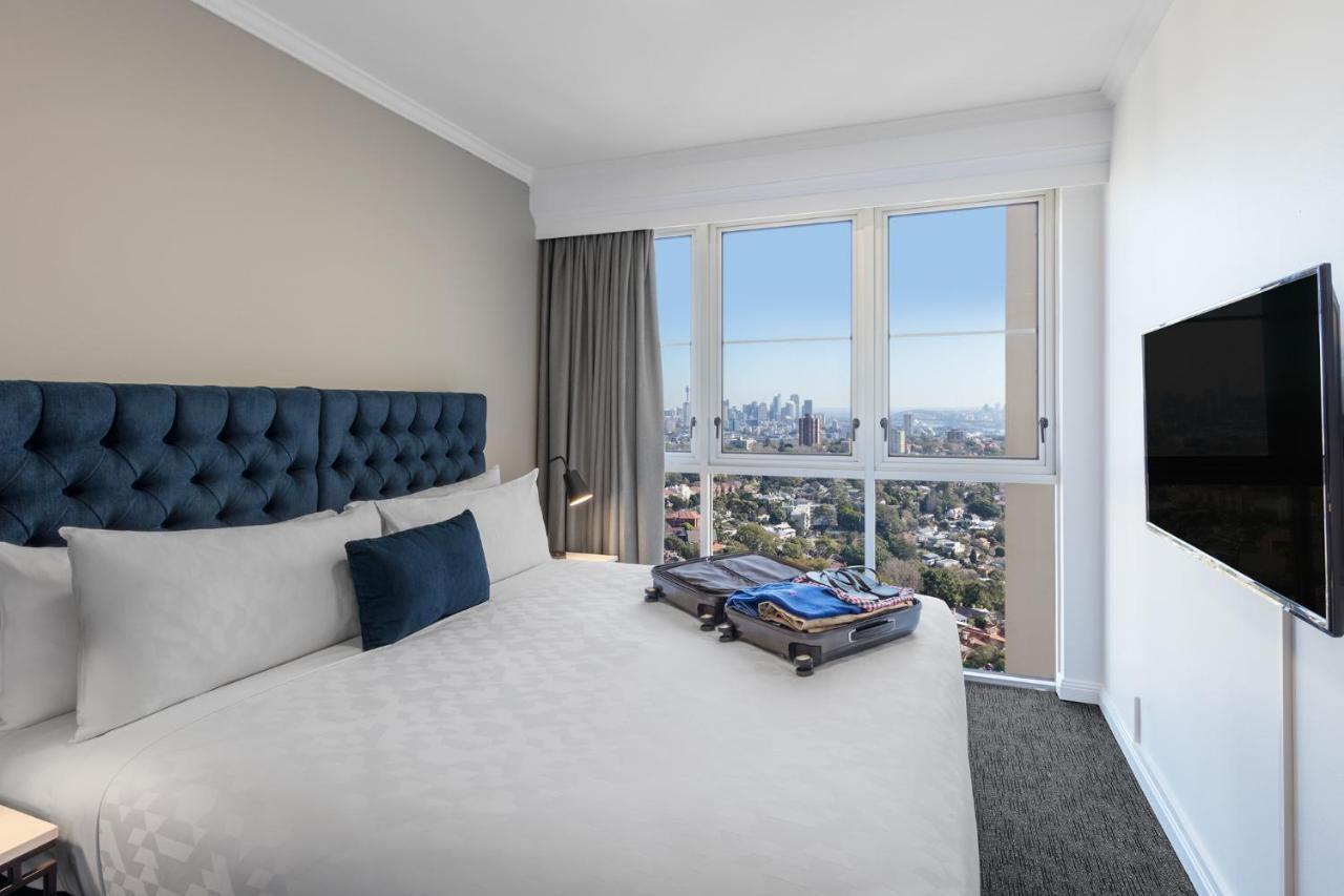 Meriton Suites Bondi Junction Sydney Exterior foto The photo shows a modern hotel bedroom featuring a king-sized bed with a blue tufted headboard. The bed is neatly made with white linens and a decorative blue pillow. There is a large window that offers a view of the city skyline, allowing natural li