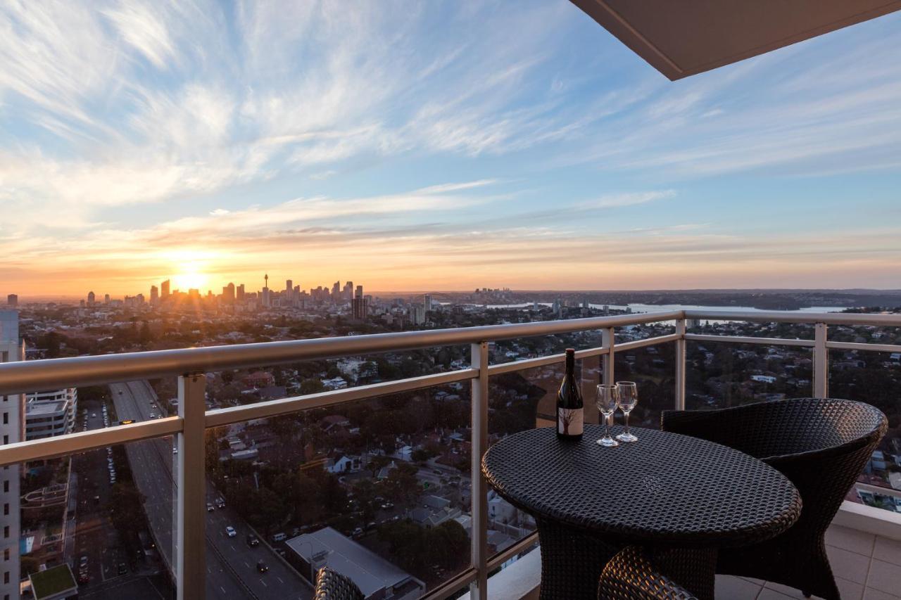 Meriton Suites Bondi Junction Sydney Exterior foto The image shows a scenic view from a balcony or terrace overlooking a cityscape during sunset. In the foreground, there is a small round table with two glasses and a bottle of wine, suggesting a romantic or relaxing setting. The sky is filled with vi