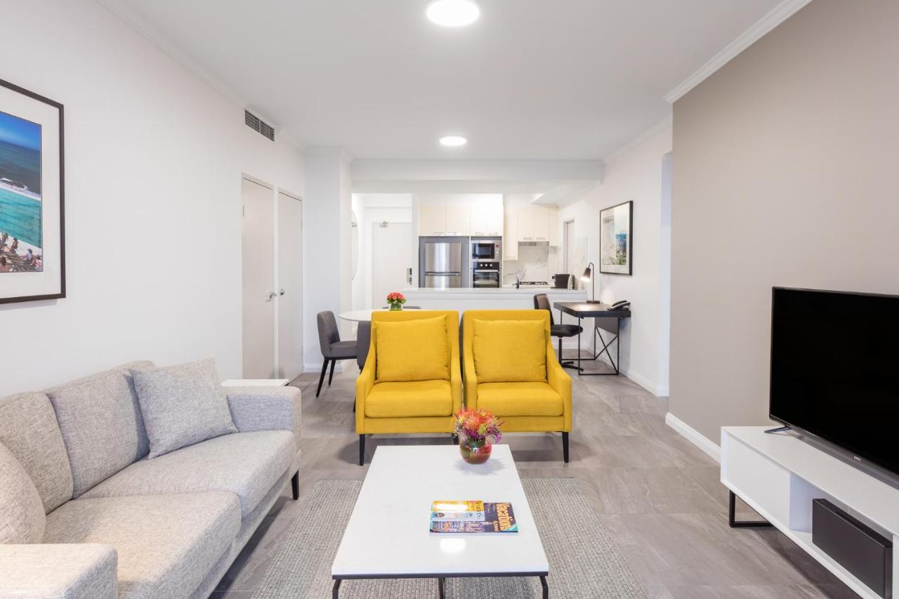 Meriton Suites Bondi Junction Sydney Exterior foto The photo shows a modern living space with a light and airy design. In the foreground, there are two bright yellow armchairs flanking a white coffee table, which has a small vase of flowers and a magazine on it. To the left, there is a light gray sof