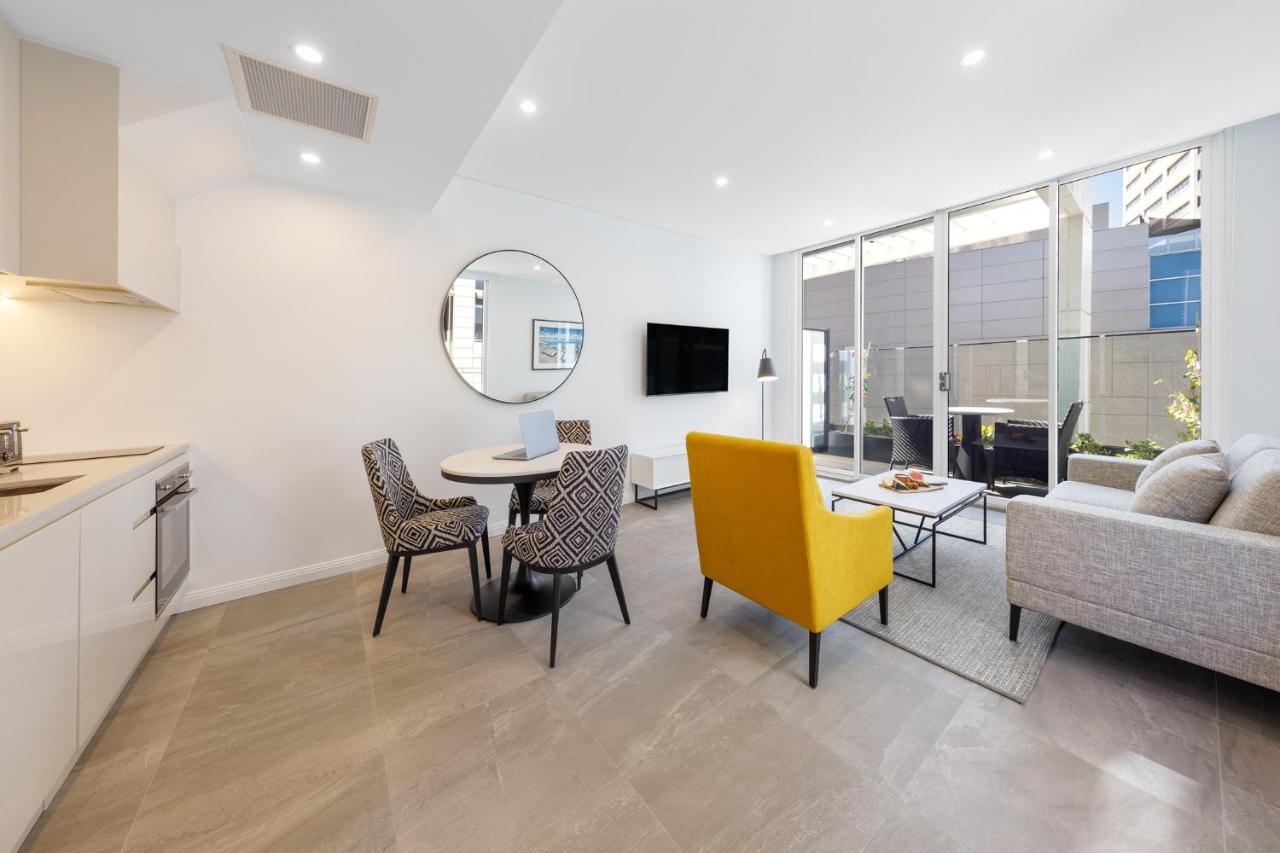 Meriton Suites Bondi Junction Sydney Exterior foto The photo shows a modern and stylish living space. On one side, there is a kitchen area with sleek cabinetry, and next to it, a round dining table surrounded by three patterned chairs. In the center of the room, there is a gray sofa accompanied by a 