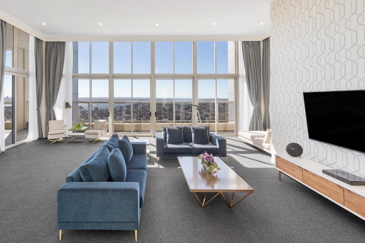Meriton Suites Bondi Junction Sydney Exterior foto The photo shows a modern and spacious living area. It features large windows that provide a panoramic view of the outside landscape and allow natural light to fill the room. The interior design includes a blue sectional sofa placed in the center, com