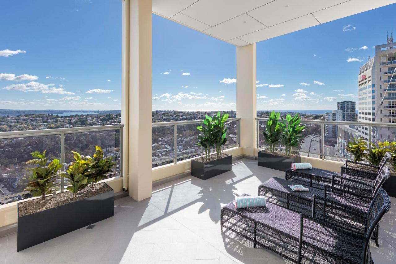 Meriton Suites Bondi Junction Sydney Exterior foto The photo shows a modern balcony or terrace area with a stunning view. There are several planters with green plants placed along the edge. The seating area features lounge chairs in a contemporary design, likely made of woven materials. The space app