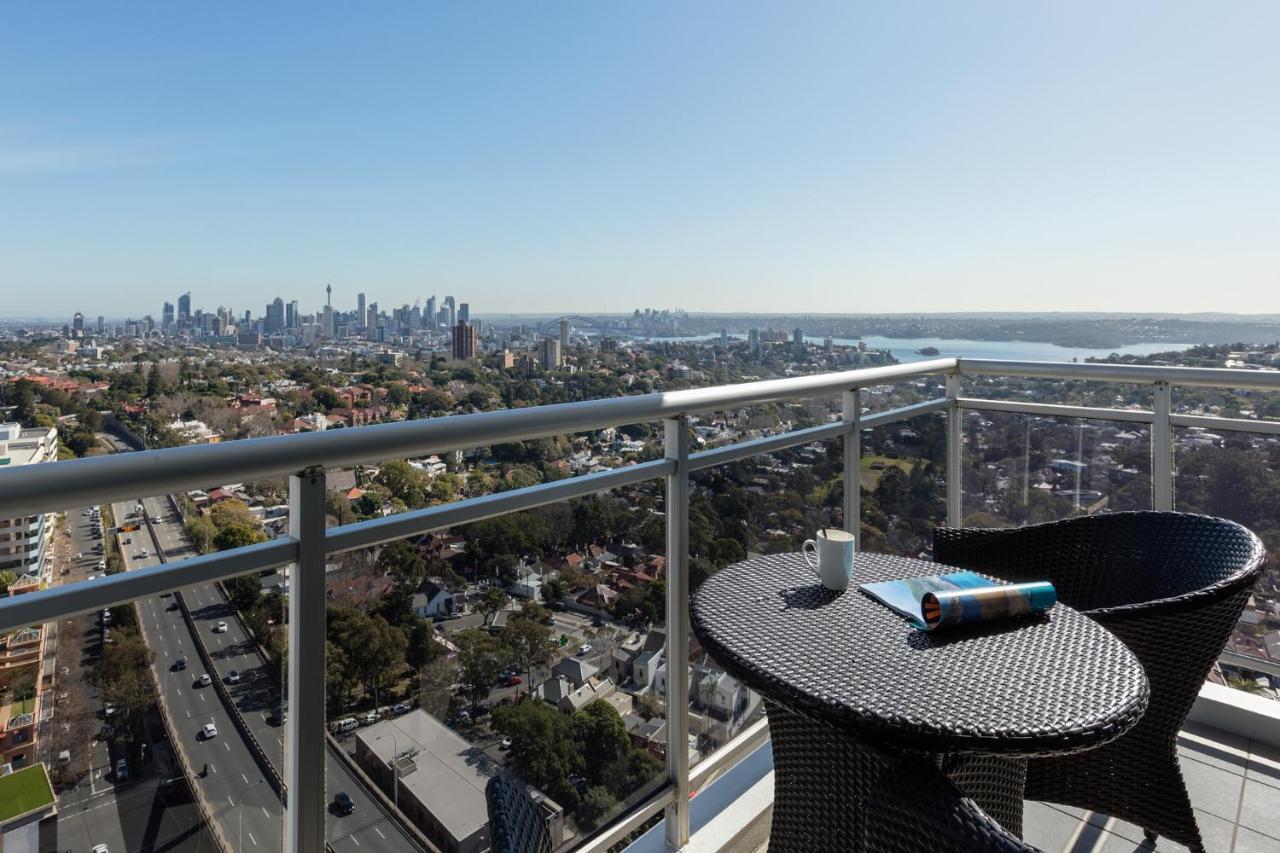 Meriton Suites Bondi Junction Sydney Exterior foto The photo shows a balcony with a view overlooking a cityscape and a body of water in the distance. There is a round table with a woven design and two chairs, one of which has a mug on it. On the table, there’s also a rolled-up magazine or document. T