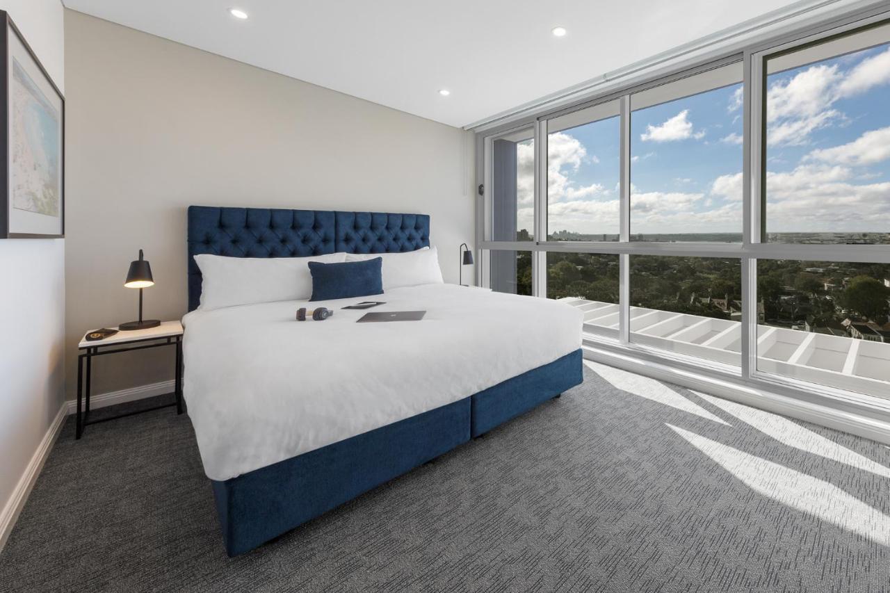 Meriton Suites Bondi Junction Sydney Exterior foto The photo shows a modern and stylish hotel room. It features a comfortable queen-size bed with a blue tufted headboard. The bed is neatly made with white linens and pillows. Next to the bed, there is a small bedside table with a desk lamp. 

On the f