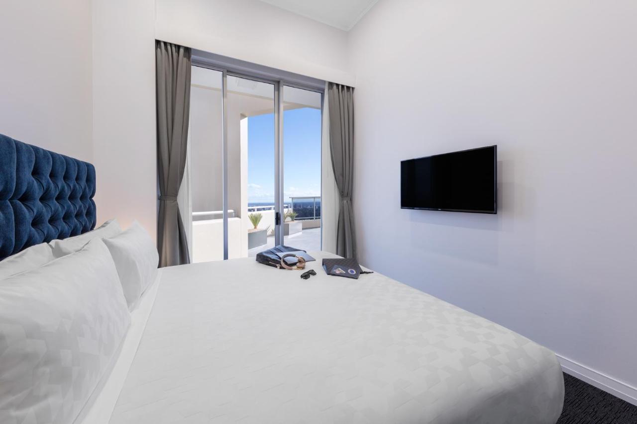 Meriton Suites Bondi Junction Sydney Exterior foto The photo shows a modern hotel room with a large, comfortable bed featuring a blue tufted headboard. There are light-colored walls, and the room is decorated simply. A flat-screen TV is mounted on the wall opposite the bed. The room has large glass d