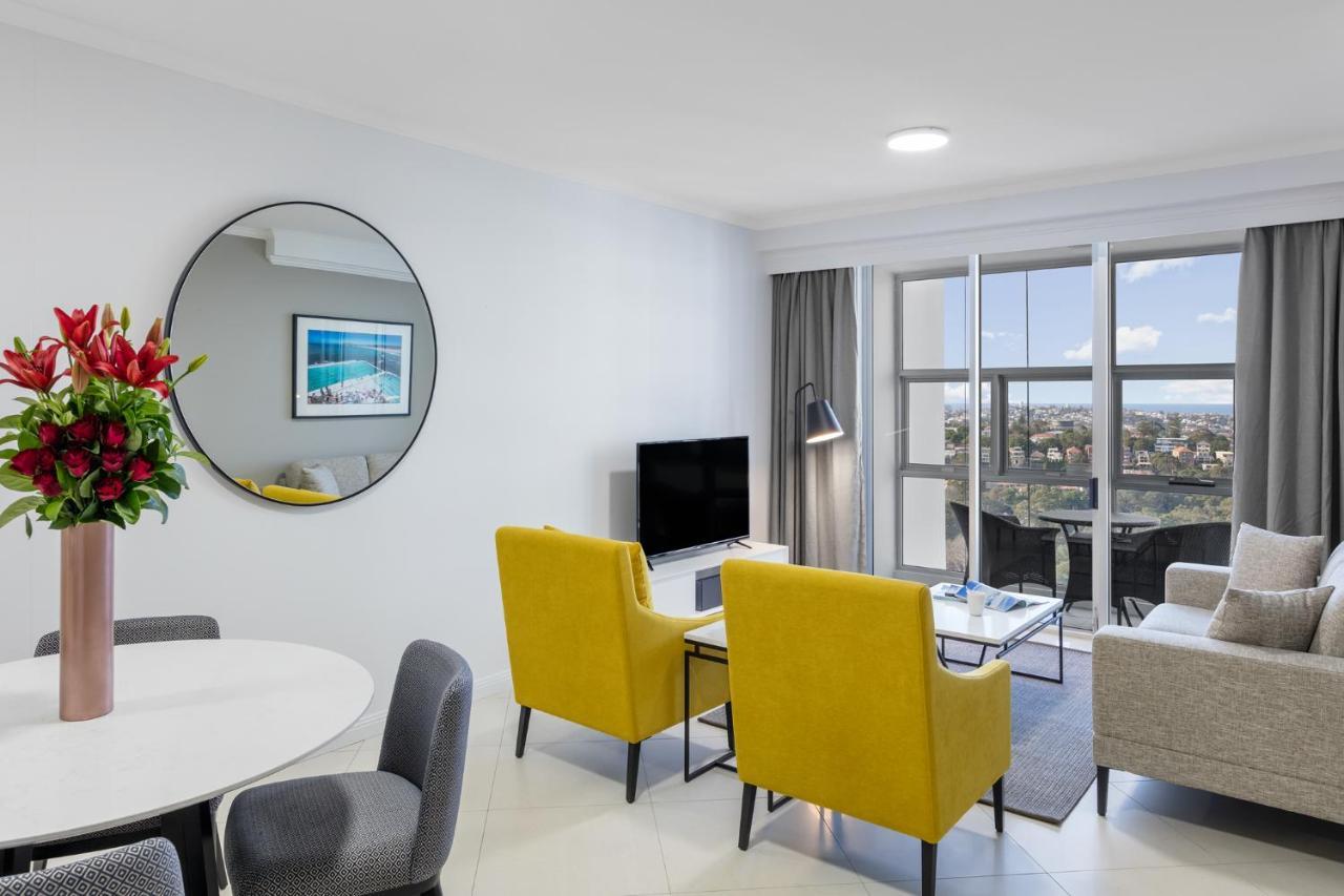 Meriton Suites Bondi Junction Sydney Exterior foto The photo shows a modern living space with a stylish design. There are two bright yellow chairs facing a small glass table, and a comfortable beige sofa to the right. A TV is positioned on the wall in front of the chairs. To the left, there's a dinin