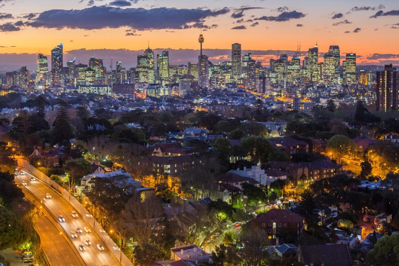 Meriton Suites Bondi Junction Sydney Exterior foto The photo captures a stunning city skyline at dusk, featuring towering skyscrapers adorned with lights against a colorful sunset backdrop. In the foreground, a green area with trees and houses is visible, while a road with cars can be seen winding th