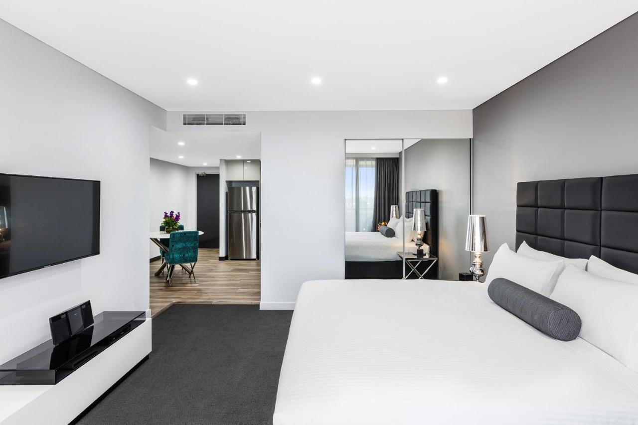 Meriton Suites Bondi Junction Sydney Exterior foto The photo shows a modern hotel room. It features a large bed with a white covering and gray accent pillows. To the left, there's a flat-screen TV mounted on the wall above a sleek white cabinet. The flooring is a light wood tone, and a large mirror i