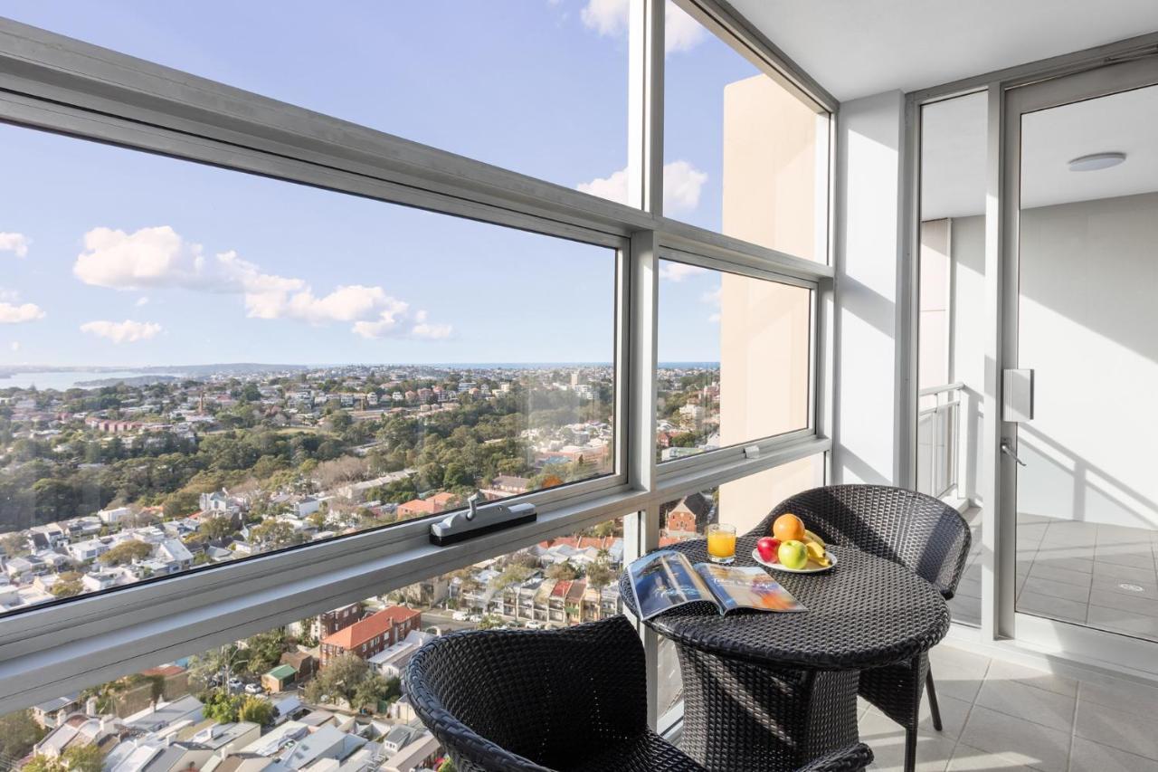 Meriton Suites Bondi Junction Sydney Exterior foto The photo shows a modern balcony with large glass windows that offer a panoramic view of a sprawling urban landscape. There is a small round table made of dark wicker, accompanied by two matching chairs. On the table, there is a bowl of fruit and a m